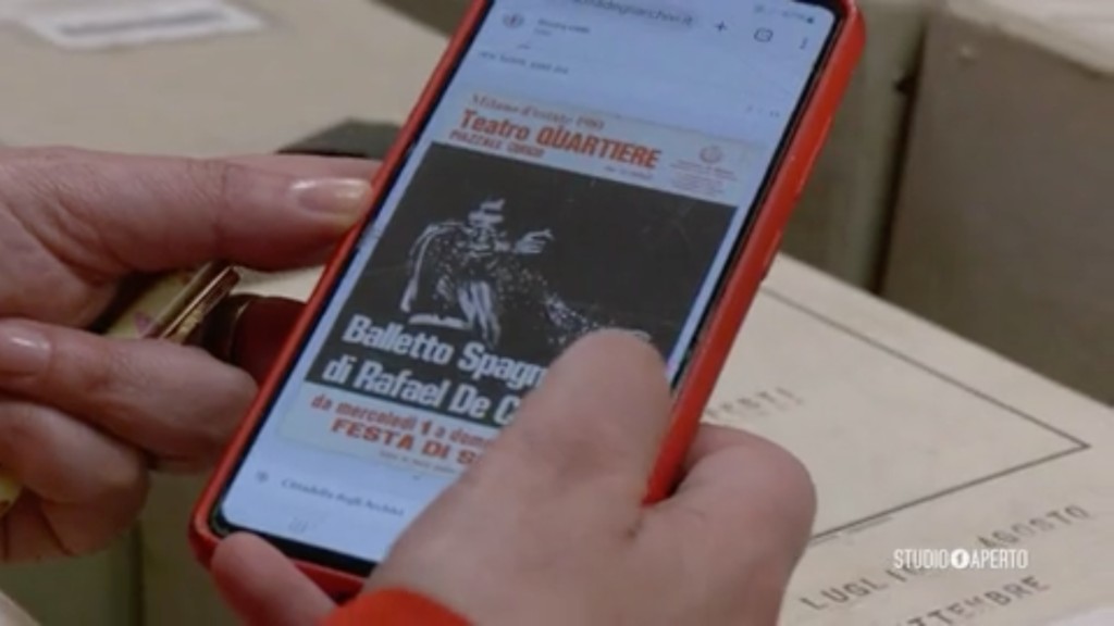 Una mano consulta i contenuti digitali di un faldone della Cittadella degli Archivi di Milano tramite tap NFC, mentre sullo schermo appare la locandina storica del Teatro Quartiere conservata nell'archivio