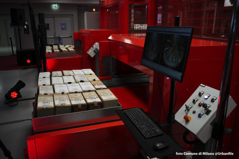 Faldoni archivistici ordinati su un piano di lavoro della Cittadella degli Archivi di Milano, accanto a una postazione operativa con monitor che mostra documenti scansionati