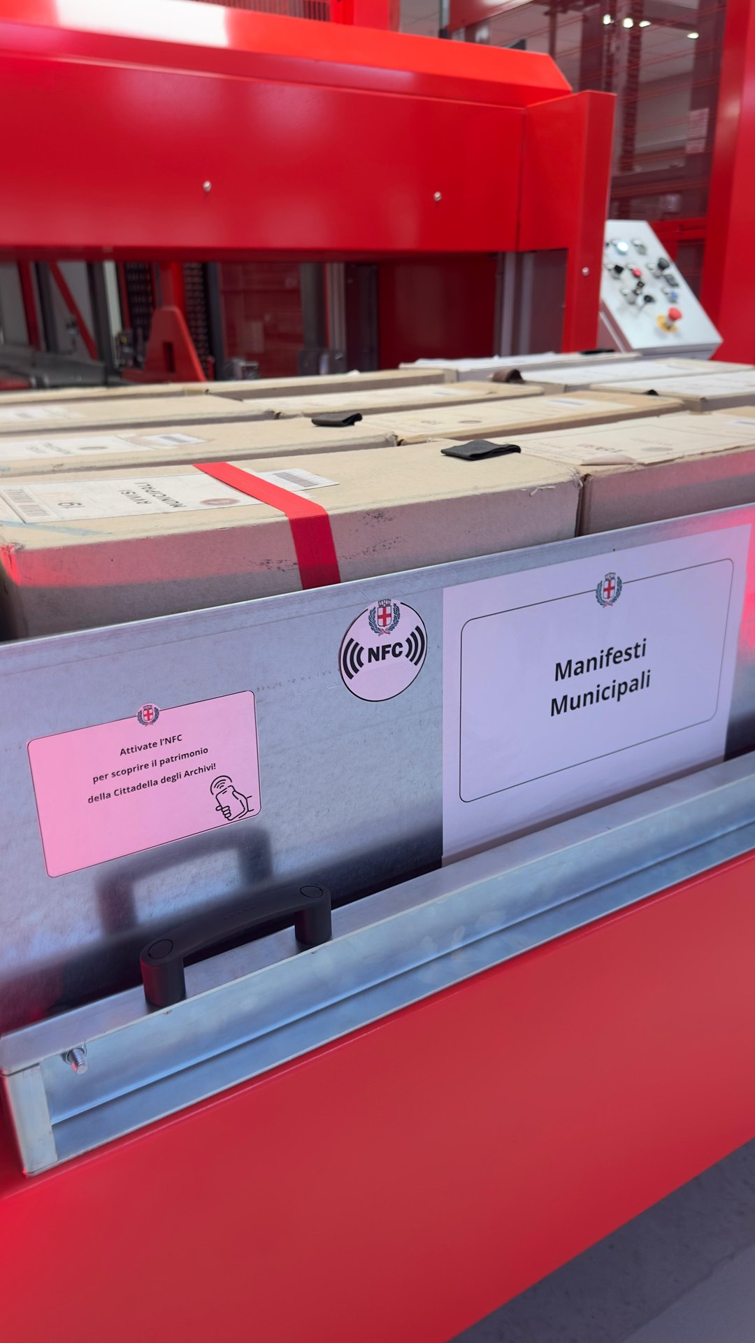 Dettaglio di un faldone della Cittadella degli Archivi etichettato Manifesti Municipali, con icona NFC e l'invito Attivate l'NFC per scoprire il patrimonio della Cittadella degli Archivi